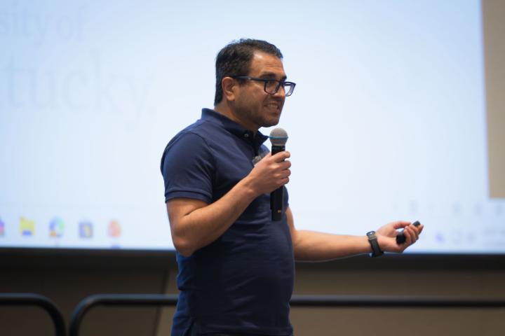 A speaker delivering a presentation from the stage, with a projector screen displaying slides behind them.