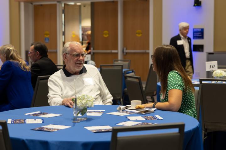 Two people talking at a round table with blue tablecloth.