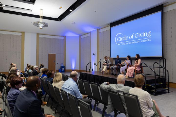 Wide view of panel on stage under “Circle of Giving” screen with audience.
