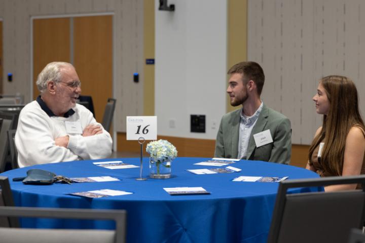 Older man in white sweater speaking with two students at round table.