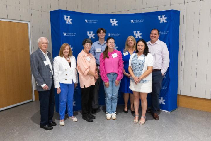 Group of eight smiling in front of UK College of Arts and Sciences step-and-repeat backdrop.