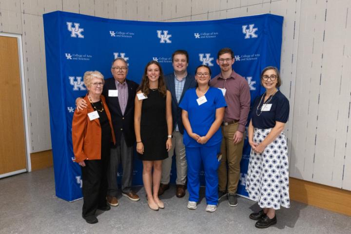 Group of seven, including student in scrubs, smiling in front of backdrop.