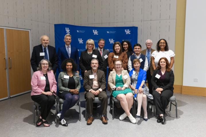 Large group of 15 seated and standing in front of UK A&S backdrop.