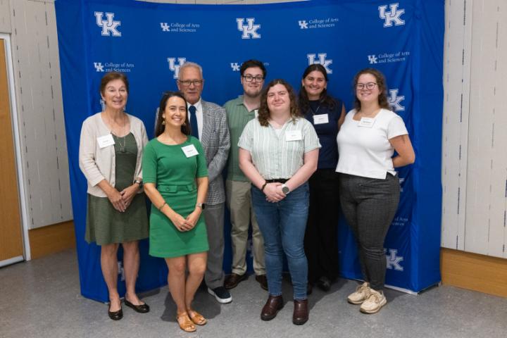 Group of seven smiling in front of UK A&S backdrop.