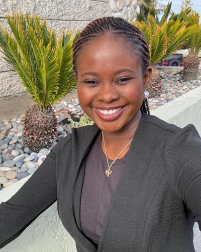 A woman in a dark blazer and necklace smiles while taking a selfie in front of palm trees and a stone wall.