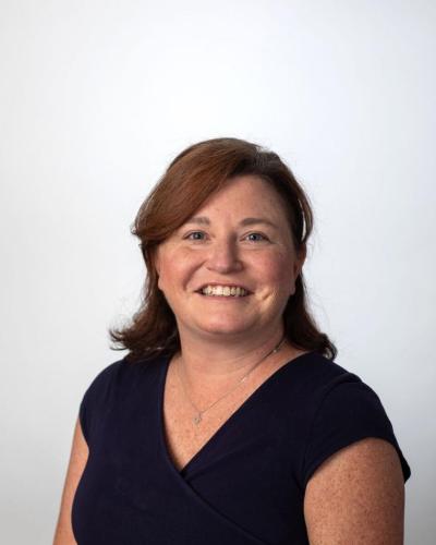 A woman with brown hair and a dark blue top smiles while wearing a necklace.
