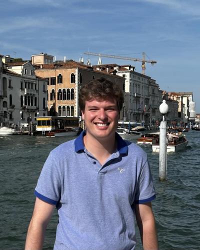A young man stands in front of a body of water with buildings and boats in the background.