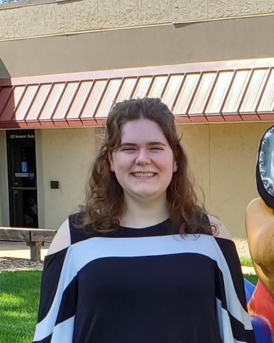 A woman with long brown hair wearing a black and white top smiles while standing outside a building.