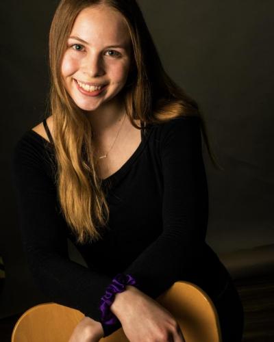 A young woman with long brown hair wearing a black outfit sits on a wooden object with her hands clasped together.