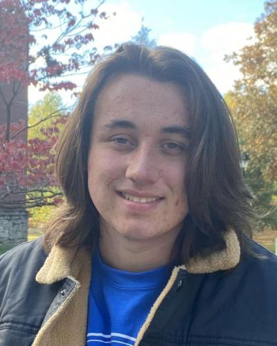 A young man with long brown hair smiling in front of trees and a building.