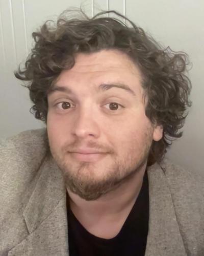 A man with curly hair and a beard, wearing a gray blazer and black shirt, looks at the camera.