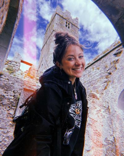 A young woman smiles while standing under an archway with a large stone building in the background.