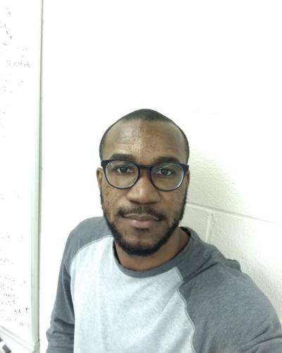 A man wearing glasses and a gray and white shirt stands in front of a white wall.