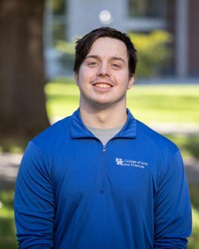 A man wearing a blue shirt with white text on it smiles while standing outside on a sunny day.