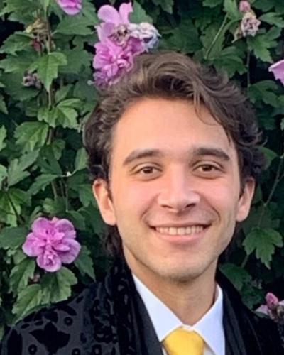 A man in a black jacket and yellow tie smiles while standing in front of a wall of greenery and purple flowers.