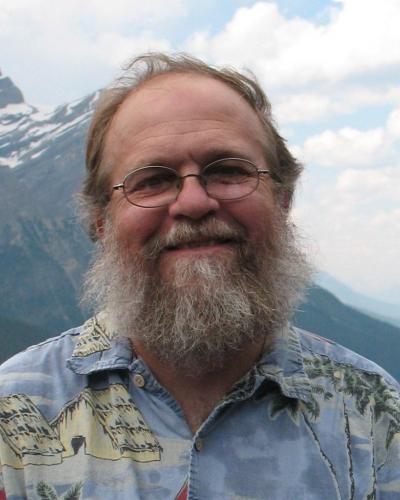 A man with a beard and glasses smiles in front of a mountainous landscape.