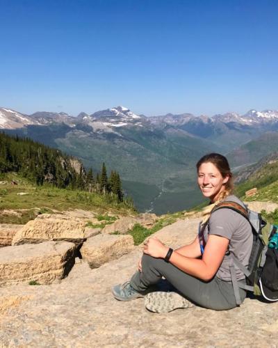 A woman sits on a rocky outcropping with a backpack, overlooking a scenic mountainous landscape.