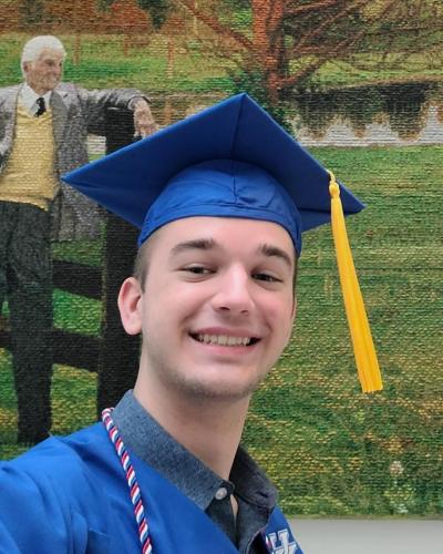 A young man in a blue graduation cap and gown smiles in front of a large photo of a man in a yellow shirt and gray jacket.