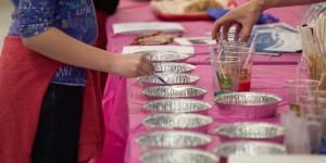 A person and an adult's hand reach for small cups on a table covered with pink paper and filled with various art supplies.