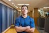 Kaleb Diaz, a University of Kentucky student, stands confidently with arms crossed in a brightly lit campus hallway. He wears a navy polo shirt and looks directly at the camera, embodying determination and leadership.