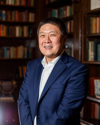 Brad Plaster smiling in a navy suit jacket, white dress shirt, and in a library