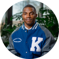 Headshot of a young man smiling, wearing a blue and gray University of Kentucky letterman jacket, with greenery visible in the background.