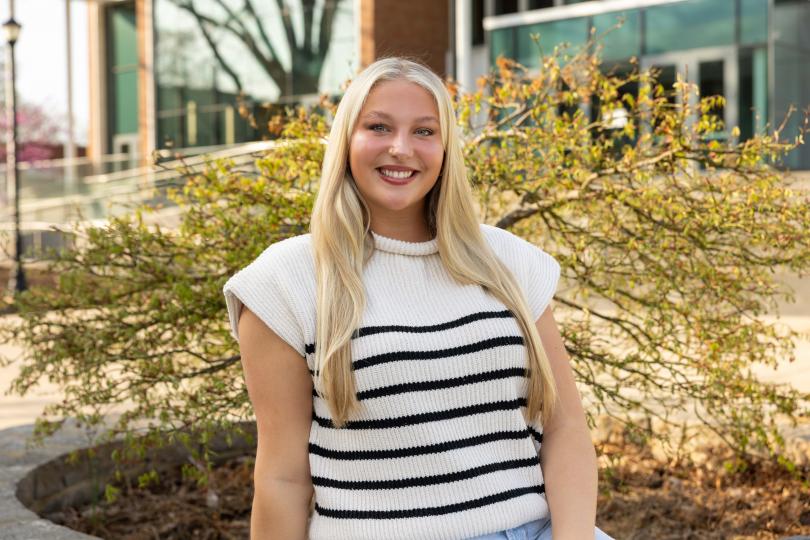 Kennedy Fitzpatrick, a smiling young woman with long blonde hair, sits outdoors in front of a budding shrub and campus buildings. She is wearing a sleeveless white knit top with black horizontal stripes.