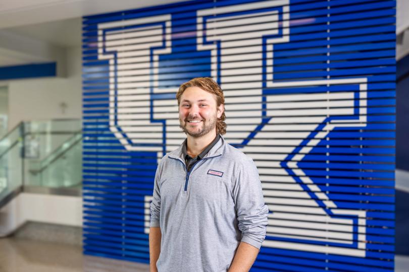 Ryson Neeley, a smiling college student with light brown hair and a trimmed beard, stands in front of a large blue and white University of Kentucky “UK” logo wall. He is wearing a light gray pullover and dark pants, posing confidently in a bright campus building.