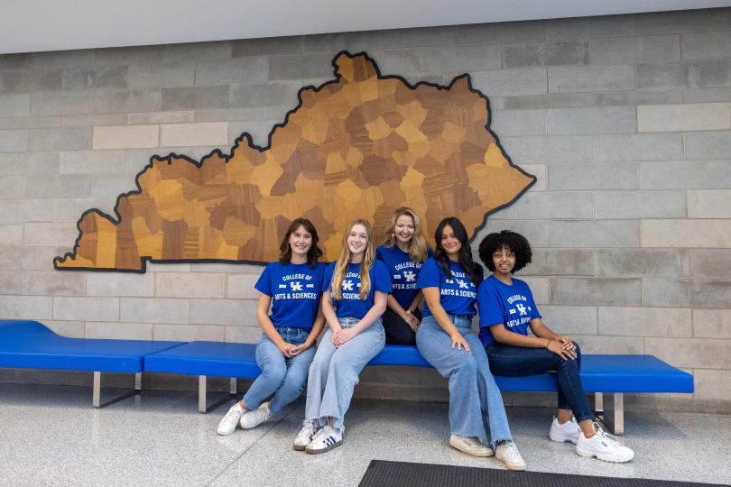 SLIPAS staff seated together on a blue bench inside a campus building, wearing matching blue College of Arts & Sciences shirts in front of a large wall mural of the state of Kentucky.